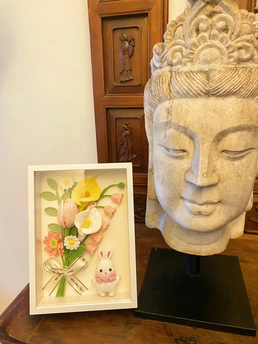 石雕旁的白色相框，内有柔和色调的钩织花朵及小兔子
