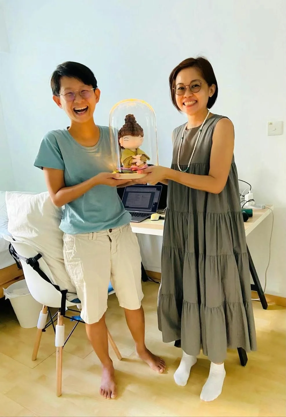 两位面带笑容的人共同捧着透明展示罩内的完成钩织玩偶